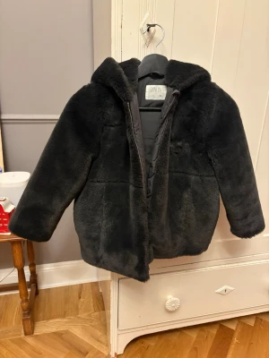 Svart pälsjacka från Zara strl 128 - Mysig svart pälsjacka från Zara med huva och dragkedja. Jackan har en fluffig känsla och är perfekt för kyliga dagar. Enkel och stilren design som passar till många outfits.