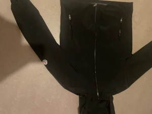 Svart vindjacka från Moncler - Svart vindjacka från Moncler med huva och hel dragkedja framtill. Jackan har två snedställda fickor med dragkedja och Moncler-logga på ena ärmen. Insidan har ett unikt serietidningsmärke. Perfekt för dig som vill ha en stilren och sportig look.