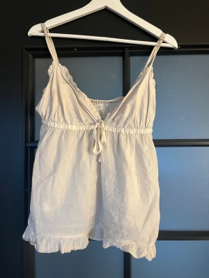 Vit linnetopp med volang från Odd Molly - Supersöt vit linnetopp från Odd Molly med justerbara axelband, volangkant nertill och broderad logga på ryggen. Toppen har knyt framtill och är gjord i mjukt bomullstyg