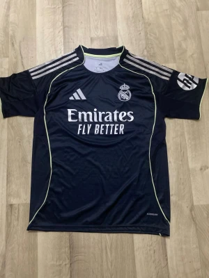 Real Madrid Adidas fotbollströja - Snygg marinblå Real Madrid matchtröja från Adidas med vita och ljusgula detaljer. Tröjan har klassiska Adidas-ränder på axlarna, klubbmärke på bröstet och 'Emirates Fly Better' tryckt framtill. Tillverkad i lätt polyester med Aeroready-teknologi.