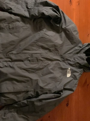 Grå  vinterjack  från The North Face - Säljer en grå vinterjacka  från The North Face med huva och vit logga på bröstet. Jackan har dragkedja framtill och är tillverkad i slitstark polyester. Perfekt för dig som gillar stilren och funktionell design. Den passar någon som är runt 160cm lång 