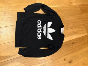 Svart Adidas sweatshirt med logga - Svart sweatshirt från Adidas med den klassiska vita treklöver-loggan och Adidas-tryck på bröstet. Tröjan har rund hals och långa ärmar, perfekt för en sportig och avslappnad stil. Passar dig som gillar streetwear och ikoniska märken.