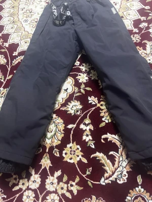 Svarta täckbyxor från Nellie - Svarta täckbyxor från Nellie med elastisk midja, justerbara hängslen och dragkedja fram. Byxorna har resår vid benslut för extra skydd mot snö och kyla. Perfekta för vintern och designade i Finland. Snygga och praktiska för kalla dagar.