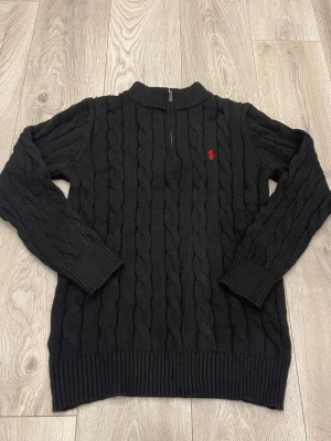 Svart stickad tröja Polo Ralph Lauren - Svart kabelstickad tröja från Polo Ralph Lauren med röd broderad logga på bröstet. Tröjan har half zip vid halsen, ribbade muddar och långärmad passform. Perfekt för dig som gillar klassisk stil med en modern twist.