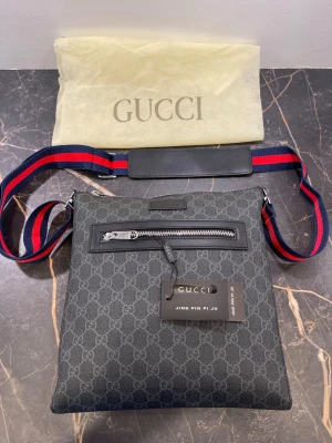 Svart axelväska från Gucci med GG-mönster - Snygg svart axelväska från Gucci med klassiskt GG-mönster över hela väskan. Justerbar axelrem i blått och rött, silverfärgad dragkedja framtill och innerfack. Väskan är rektangulär och har detaljer i skinn. Perfekt för dig som vill ha en stilren och lyxig look.