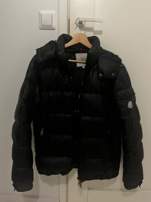 Svart Moncler pufferjacka herr - Svart pufferjacka från Moncler med klassisk logga på ärmen och snygga detaljer som ficka med knapp. Jackan har dragkedja framtill och är tillverkad i glansig syntet med dunfyllning. Perfekt för kalla dagar och har en modern, lite loose passform.