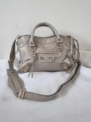 Grå/beige guld väska vintage style y2k 2000s - Snygg grå axelväska i skinnimitation med guldfärgade metalldetaljer och dragkedja. Väskan har både handtag och justerbar axelrem, samt flera fack och dekorativa nitar framtill. Perfekt storlek för att få plats med allt du behöver.