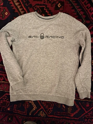 Grå Sail Racing sweatshirt - Snygg grå sweatshirt från Sail Racing med svart logotyp tryckt på bröstet och en mindre logga i nacken. Tröjan har rund hals, ribbade muddar och är tillverkad i mjuk bomullsmix. Perfekt för chill dagar och enkel att matcha med jeans eller joggers.