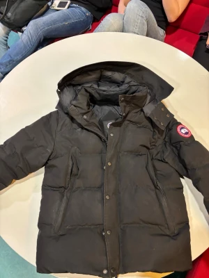 Svart dunjacka från Canada Goose - Säljer en svart dunjacka från Canada Goose med ikonisk patch på ärmen och stor huva. Jackan har dragkedja och knappar framtill, två stora fickor och quiltad design. Perfekt för kalla dagar och riktigt snygg till vinterlooken.