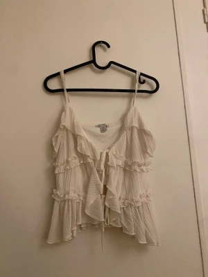 Vit omlott linnetopp med volanger - Supersöt vit linnetopp från newyorker med tunna axelband, omlott-design och volanger. Toppen har knyt framtill och är tillverkad i ett lätt, ribbat material med diskreta ränder. Perfekt för sommarens alla häng och selfies!
