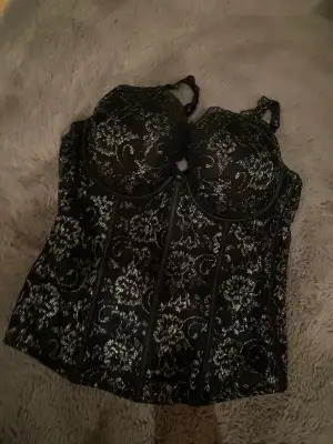 Säljer en snygg korsett i svart med glittriga silverdetaljer och blommigt mönster. Toppen har formade kupor, justerbara axelband och markerade sömmar som ger en cool siluett. Perfekt för dig som vill sticka ut med lite extra bling och glam. Och parfeckt för nyår.