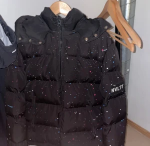 Svart pufferjacka från NVLTY med färgstänk - Svart pufferjacka från NVLTY med coola färgstänk i blått, vitt och rosa. Jackan har huva, dragkedja framtill och en patch med NVLTY på ärmen. Perfekt för dig som gillar streetstyle och vill sticka ut lite extra under kalla dagar. Kom dm för mer bilder! Pris går att diskuteras!