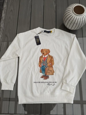 Vit sweatshirt Polo Bear Ralph Lauren - Vit sweatshirt från Polo Ralph Lauren med ikoniskt Polo Bear-tryck framtill. Tröjan har rund hals, ribbade muddar och är tillverkad i 100% bomull. Perfekt för dig som gillar klassisk stil med en lekfull twist.