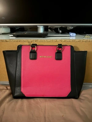 Svart och rosa handväska Michael Kors - Snygg handväska från Michael Kors i svart och starkt rosa med guldfärgade metalldetaljer. Väskan har två handtag, dragkedja upptill och rymligt foder med logomönster. Perfekt för dig som vill sticka ut med en färgklick och ändå ha plats för allt du behöver.