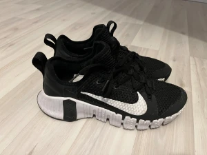 Svarta Nike Metcon sneakers - Nike Metcon sneakers i svart med vit sula och vit swoosh på sidan. Ovandelen är i ventilerande mesh och har snörning för bra passform. Sulan är robust och designad för stabilitet vid träning. Skorna har rund tå och platt sula, perfekt för gym och styrketräning. 3 månad gammal 