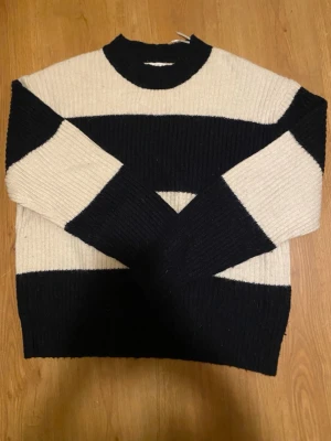 Randig stickad tröja i marinblå och vit - Säljer en ribbad stickad tröja med breda svarta och beige ränder. Tröjan har rund hals och långärmade ärmar, perfekt för dig som gillar klassisk stil med en twist. Passar dig som vill ha en mysig och snygg look.
