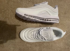Nike Air Max 97 helvita sneakers - Nike Air Max 97 sneakers i helvit färg med ikoniska vågiga linjer och synlig Air-sula. Skorna har snörning, rund tå och platt sula. Ovandelen är i syntet och mesh för skön komfort och cool look. Perfekta för dig som vill ha en clean och sportig stil.