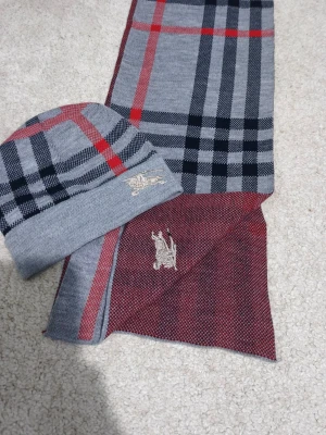 Grå rutig bomullsväst med broderi - Snygg väst från Burberry i 100% bomull med klassiskt rutigt mönster i grått, svart och rött. Västen har ett broderat Burberry-ryttaremotiv framtill och en stilren, rak form. Perfekt för dig som gillar ikoniska mönster och vill sticka ut med en cool detalj.