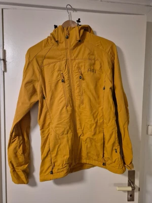 Senapsgul vindjacka från P4H med huva - Snygg vindjacka från P4H med huva och flera dragkedjeförsedda fickor. Jackan har en sportig look, lång ärm och är tillverkad i slitstark bomull. Perfekt för dig som gillar friluftsliv och vill sticka ut med färg. Designad i Sverige. Ett litet hål i innerfickan, syns på ena bilden. 