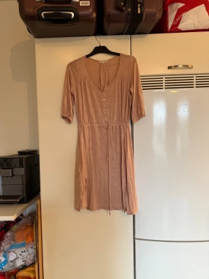 Beige kortklänning med knappar - Supersöt gamelrosa klänning med knappar framtill, knytband i midjan och trekvartsärm. Klänningen har diskret paisleymönster i tyget och är gjord i mjuk bomull. Perfekt för dig som gillar en avslappnad men ändå stilren look. Från märket Odd Molly. Storlek 1 som enligt deras storleksguide motsvarar en S. men ör mer en M. Fint skick, men tyvärr saknas första knappen. 
