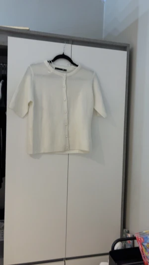 Vit kofta med pärlknappar från 157 XS - Supersöt vit kofta från 157 i storlek XS. Den har korta ärmar och pärlknappar framtill som ger en extra touch. Koftan är stickad och har en mjuk, ribbad kant vid halsen. Perfekt att slänga på sig när det är lite kyligt ute.