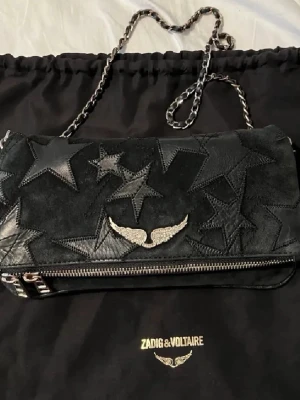 Svart axelväska med stjärnor från Zadig & Voltaire - Limited edition svart axelväska från Zadig & Voltaire med stjärnmönster i mocka och skinn. Väskan har en silverfärgad kedja, dragkedja och vinge. Dustbag följer med och en kort och en lång kedja. Skriv om du har frågor💕köpt för 3000kr. Priset kan diskuteras