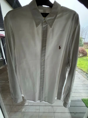 Vit skjorta från Ralph Lauren XXL - Vit skjorta från Ralph Lauren i storlek XXL med klassisk krage och knappar framtill. Slim fit-modell med det ikoniska broderade Polo-logot på bröstet. Skjortan har långa ärmar och är tillverkad i mjuk bomull som känns skön mot huden.
