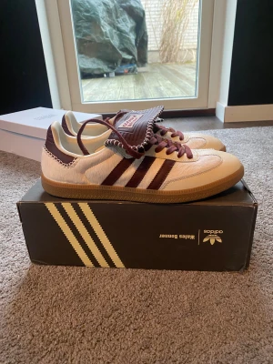 Adidas Wales Bonner Samba sneakers - Unika Adidas Wales Bonner Samba sneakers i beige och vinröd med klassiska ränder och kontrasterande snörning. Skorna har detaljerad vit stickning runt kanterna och en gummisula. Materialmix av skinn och syntet ger en lyxig känsla och retrovibe.