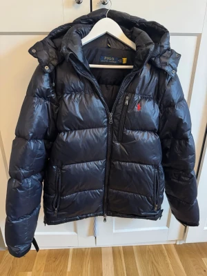 Marinblå pufferjacka Polo Ralph Lauren M - Snygg marinblå pufferjacka från Polo Ralph Lauren med huva och dragkedjor framtill. Hel och i fint skick när man tittar på jackan. Pyttelitet hål under luvan, mindre än ett knapphål. Höger ärm, litet hål samt på mudden (se bild). Köpt på Hede outlet i Kungsbacka för ett år sedan. För liten för sonen nu därav försäljningen.