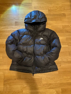 Svart dunjacka från Nike - Svart pufferjacka från Nike i storlek Xl junior passar 158-170 med Therma-Fit teknologi. Jackan har huva, dragkedja framtill och diskret Nike-logga på bröstet. Fylld med syntetmaterial för extra värme och har långa ärmar med elastiska muddar. Perfekt för kalla dagar
