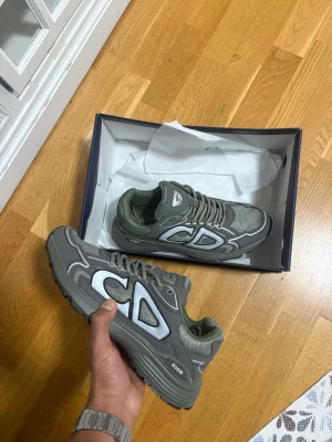 Gröna Dior B30 sneakers storlek 42 - Säljer ett par Dior B30 sneakers i en snygg olivgrön färg med grå och vita detaljer. Skorna har en chunky platt sula, mesh och syntetpaneler, samt tydlig CD-logga på sidan och hälen. Perfekt för dig som vill sticka ut med exklusiva sneakers. Pris är ej hugget i sten!