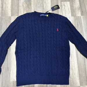 Marinblå kabelstickad tröja Polo Ralph Lauren - Kabelstickad marinblå tröja från Polo Ralph Lauren med rund hals och röd broderad logga på bröstet. Tröjan har ribbade muddar och är tillverkad i mjuk bomull. Perfekt för dig som gillar klassisk och stilren look.