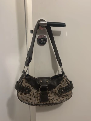 Beige och brun axelväska med mönster - Snygg axelväska i beige tyg med svart grafiskt mönster och detaljer i mörkbrunt skinn. Väskan har bred axelrem, dragkedja upptill och coola metalldetaljer. Slit på metallen visas i sista bilden!! Köpt från urban outfitters