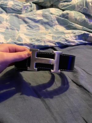 Svart skinnbälte med H-spänne - Snyggt svart bälte i skinn med ett stort, silverfärgat H-spänne. Bältet har vita sömmar längs kanterna som ger en schysst kontrast. Perfekt accessoar för att lyfta din outfit med en stilren och modern touch.