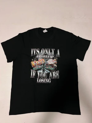 ”Its only a problem if you are losing” T shirt  - En svart t-shirt med ett stort casinoinspirerat tryck och texten “It’s only a problem if you are losing”. Motivet visar spelautomater, tärningar och ett roulettebord i färgstarka detaljer. Perfekt för den som gillar humoristiska tröjor eller casinotema. Tröjan är i nytt skick och sitter bekvämt.