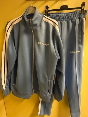 Blå Palm Angels tracksuit  - Snygg blå tracksuit från Palm Angels med vita  ränder längs axlarna. Perfekt för en chill och trendig look.