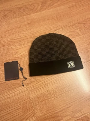 Rutig svart mössa - Snygg svart mössa med klassiskt rutigt mönster och uppvikt kant med logga framtill. Mössan har en stilren design och är tillverkad i ett mjukt material som känns lyxigt. Perfekt för dig som vill ha en exklusiv accessoar.