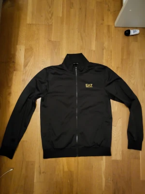 Svart EA7 ziptröja med gul logga - Svart ziptröja från EA7 med gul logga på bröstet och coola gula detaljer i nacken. Tröjan har hel dragkedja, hög krage och långa ärmar. Perfekt för en sportig och stilren look. Mycket gott skick, känns som ny. 