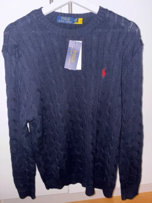 Marinblå stickad tröja Polo Ralph Lauren - Snygg marinblå stickad tröja från Polo Ralph Lauren med klassiskt kabelstickat mönster och röd broderad logga på bröstet. Rund hals och långa ärmar, perfekt för dig som gillar stilren och tidlös design. Osäker på äkhet fick den i present
