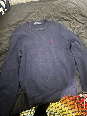 Mörkblå stickad tröja från Polo Ralph Lauren - Säljer en klassisk mörkblå stickad tröja från Polo Ralph Lauren med rund halsringning och röd broderad logga på bröstet. Tröjan har ribbade muddar vid ärmslut och nederkant, perfekt för lager-på-lager under hösten och vintern. Använd några gånger annars nästan ny