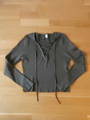 Olivgrön topp - Snygg olivgrön ribbad topp från med långa ärmar och snygg snörning framtill. Materialet är mjukt och stretchigt, perfekt för en chill men ändå trendig look. Kan matchas till både jeans och kjol.