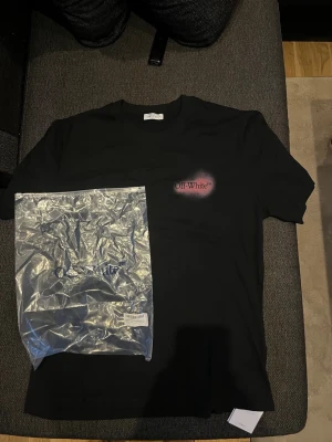 Svart Off-White t-shirt med rosa tryck - Säljer en svart t-shirt från Off-White med ikoniskt rosa sprayat tryck på bröstet och stor pilgrafik på ryggen. Klassisk passform och rund hals. Tillverkad i mjuk bomull, perfekt för dig som gillar streetwear och vill sticka ut.