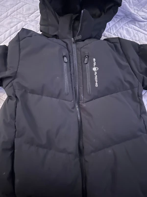 Svart pufferjacka Sail Racing 176 - Svart pufferjacka från Sail Racing med huva och två vertikala bröstfickor med dragkedja. Jackan har Sail Racing-logga på bröstet och en patch på ärmen med 'Sail Antarctica Expedition'. Perfekt för kalla dagar och har en clean, sportig look.