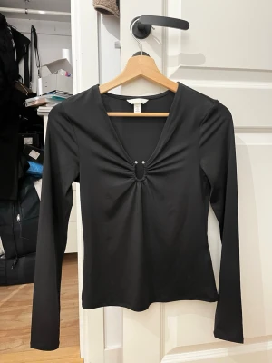 Svart långärmad topp från H&M - Svart långärmad topp från H&M i stretchigt syntetmaterial. Toppen har en djup v-ringning med snygg rynkning och två silvriga detaljer framtill. Passformen är slim och tyget är slätt och mjukt. Perfekt för dig som gillar stilrena och trendiga plagg.
