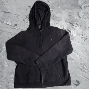 Blå  hoodie från Polo Ralph Lauren  - Blå hoodie från Polo Ralph Lauren. Tröjan har huva, dragkedja framtill och två fickor. Materialet är mjuk bomull med insida i fleece. Klassisk röd Polo-logga på bröstet och långärmad passform. Perfekt för chill dagar och streetwear.