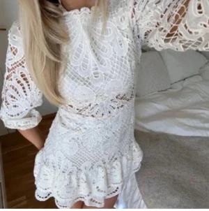 Vit spetsklänning med volang - Superfin vit kort spetsklänning med höghalsad krage, långa puffärmar och volangkant nertill. Klänningen har ett detaljerat broderat mönster och markerad midja. Perfekt för dig som vill sticka ut med en romantisk och trendig look.