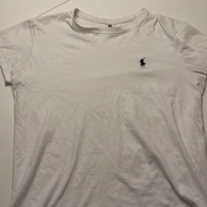 Vit t-shirt från Roly i XS - Vit t-shirt från Roly i storlek XS. Klassisk rund hals och korta ärmar, med en liten svart broderad logga på bröstet. Mjuk och skön i bomull, perfekt för en clean och enkel look.