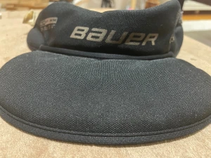 Svart hockeyhalskydd från Bauer - Säljer ett svart hockeyhalskydd från Bauer med broderad logga i grått. Skyddet har en rundad form och är tillverkat i slitstarkt syntetmaterial med mjuk insida. Justerbar rem med metallspänne för enkel passform. Perfekt för hockeyträning eller match.