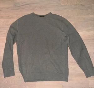 Grå stickad tröja från Dressmann - Säljer en enkel och snygg grå stickad tröja från Dressmann i storlek L. Tröjan har rund hals, diskret strukturmönster och långa ärmar med ribbade muddar. Perfekt för dig som gillar stilrena och bekväma plagg.