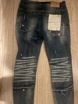 Purple Brand skinny jeans med slitningar - Säljer ett par snygga skinny jeans från Purple Brand i mörkblå tvätt med slitningar, patchade detaljer och färgstänk. Jeansen har klassisk femficksdesign, låg midja och coola kontrastdetaljer som bandana i bakfickan. Perfekt för dig som gillar streetstyle. Kommer med original box.